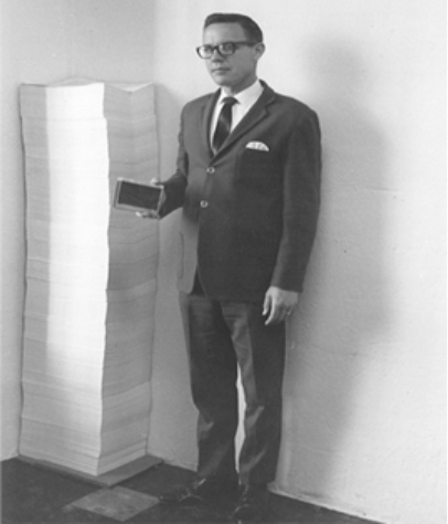 Black and white photographer of David Mulligan standing beside a stack of papers for Microfiche Publishers, Inc. in 1975
